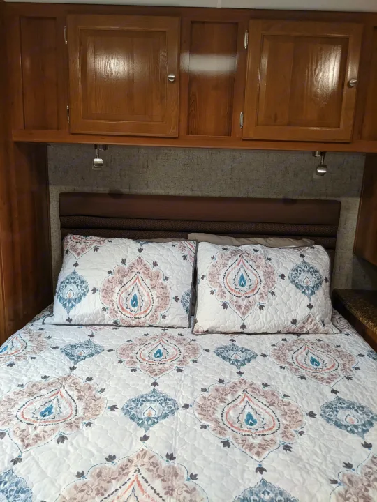 RV rental bedroom featuring a bed with patterned bedding, a headboard, wooden overhead storage, and reading lights.. Coachmen Leprechaun 2018