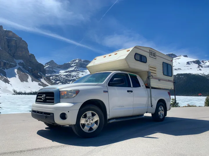 White truck with RV rental camper parked by a frozen lake, backed by snow-capped mountains under a clear blue sky. Adventure awaits!. Glacier Campers Truck Camper 2016