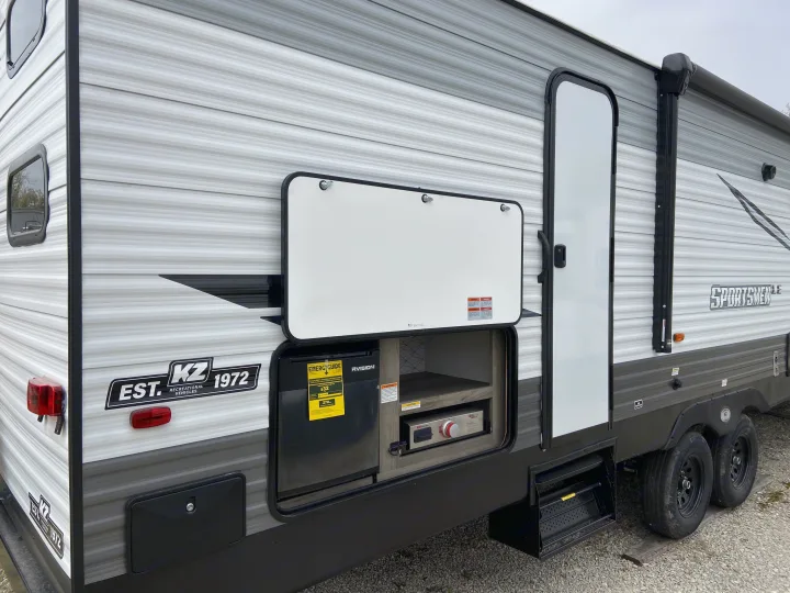 Side view of a grey and white RV, revealing an open outdoor kitchen with a mini-fridge and stove. Ideal for your next RV rental adventure.. Sportsmen LE 2021