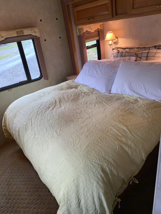 Cozy RV rental bedroom with a cream-colored bed, white and plaid pillows, and windows looking outside.. Tioga M-30u Ford 2005