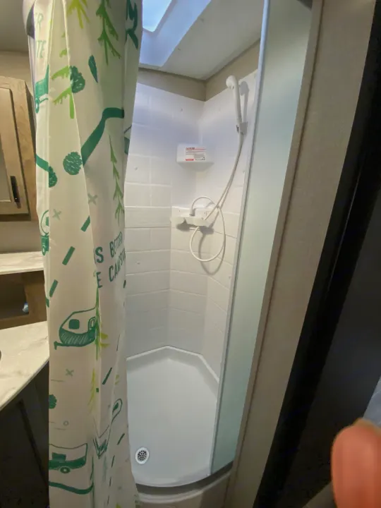 An RV rental bathroom showing a compact white shower stall with a green-patterned curtain, handheld shower, and skylight.. Sonic Lite 2021