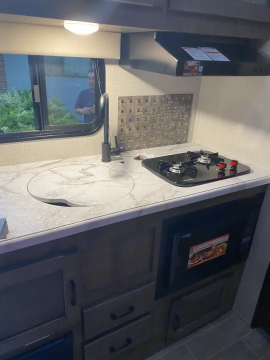 RV rental kitchen. Light counter, covered sink, 2-burner stovetop, microwave below. Dark cabinets. Reflection of a person in the window.. Sonic Lite 2021