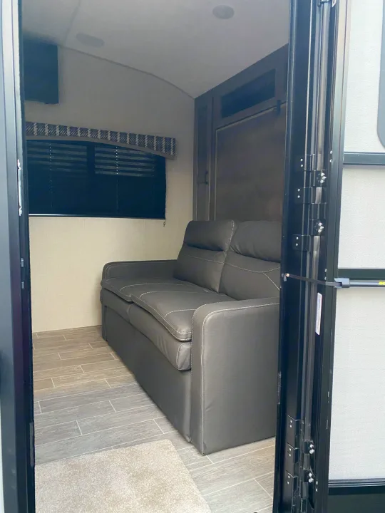Interior view of an RV rental showing a gray sofa, wall-mounted TV, window with blind, and wood-look tile floor.. Sonic Lite 2021