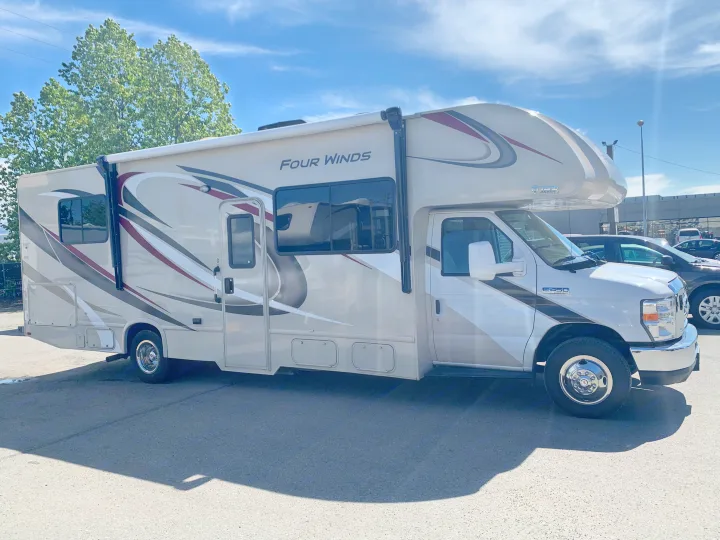 This beige Four Winds Class C rv rental motorhome, with gray & red graphics, is parked on asphalt under the sun.. Thor Motor Coach Four Winds 2019