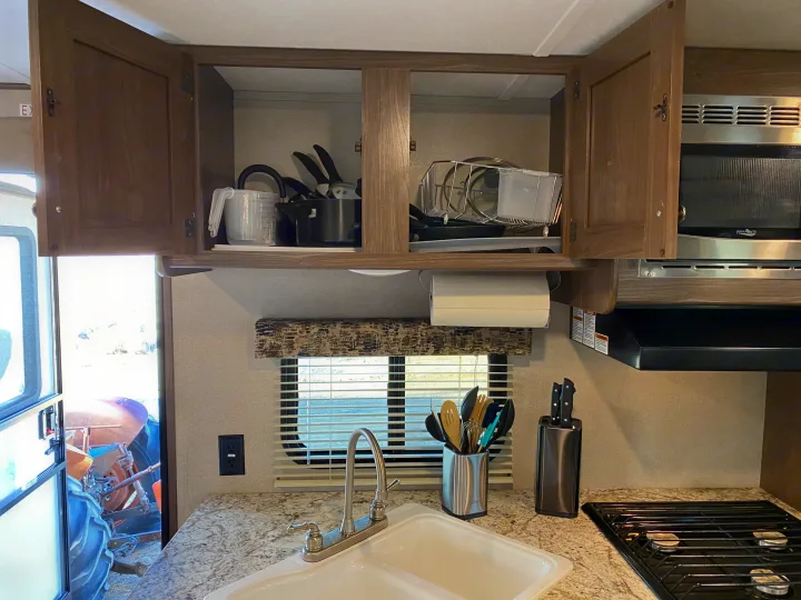 RV rental kitchen view: open cabinets, microwave, sink, stove, and an outdoor view of a tractor.. Keystone RV Hideout 2019
