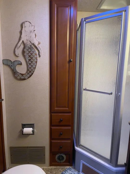 Inside an RV rental, this bathroom has a toilet, a textured glass shower, and a narrow cabinet with mermaid decor.. Other Other 2010