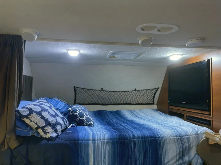 Cozy blue RV rental bedroom with striped comforter, pineapple pillows, TV, and overhead lights.. Other Other 2010