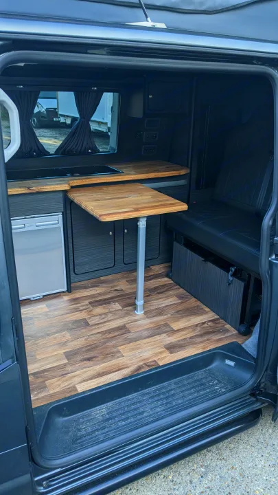View inside an RV rental camper van, showing wood flooring, a central table, fridge, and dark cabinetry. Ready for adventure.. vw T6 2016