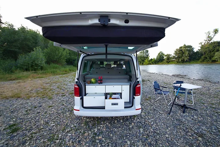 White RV rental camper van open at the back, revealing a kitchen setup, parked on a riverbank for outdoor leisure.. Volkswagen California Beach T6.1 2023