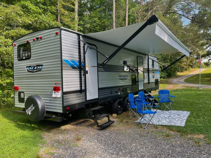 This large grey RV rental is set up at a campsite with an extended awning, blue chairs, and a patterned mat.. Forest River Salem 2021