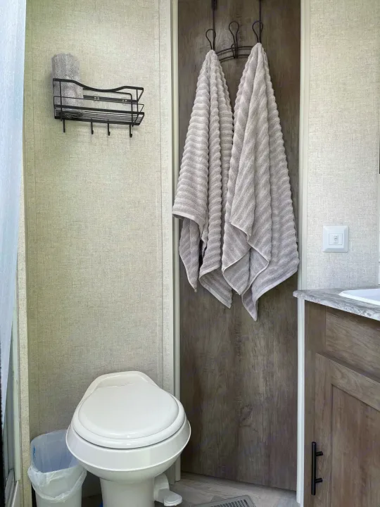 An RV rental bathroom features a toilet, waste bin, wall shelf with towel, and towels hanging on a dark wood door.. Forest River Salem 2021
