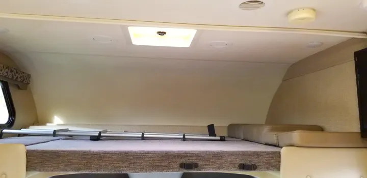 Inside an RV rental: a cozy cab-over bunk bed with overhead lighting, a window, and neutral wall textures.. Mercedes Benz Four Winds 2018