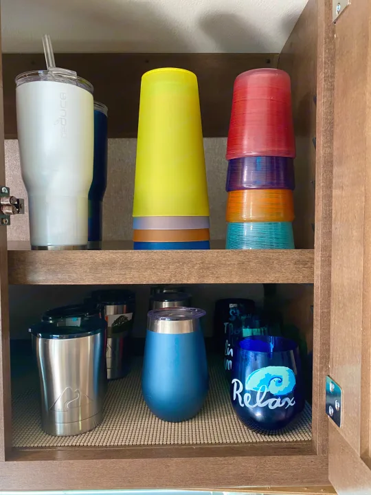 Organized RV rental cabinet with colorful stacked cups and various drinkware, including tumblers, on non-slip mats.. Winnebago Porto 2020