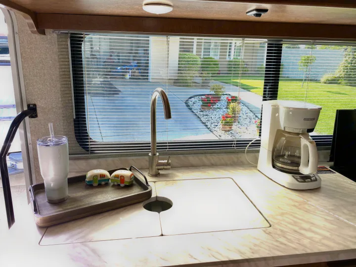 RV rental kitchen with modern faucet, Black+Decker coffee maker, and camper shakers on a light counter, overlooking a yard.. Winnebago Porto 2020