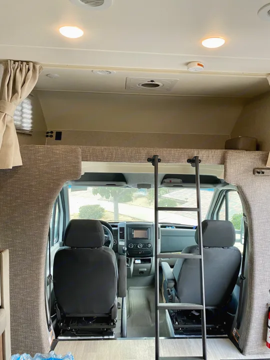 View inside an RV rental, featuring the driver's cab, dashboard, and the cozy over-cab bunk bed with a ladder.. Winnebago Porto 2020