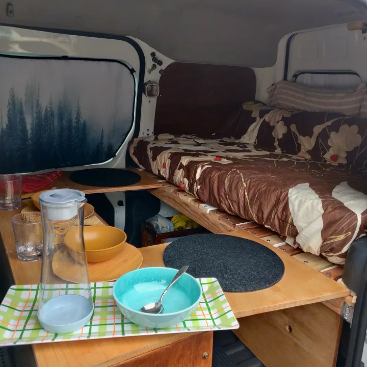 Cozy RV rental interior featuring a bed, built-in kitchen storage with bowls and glasses, and a forest-themed window shade.. Ford Connect 2012