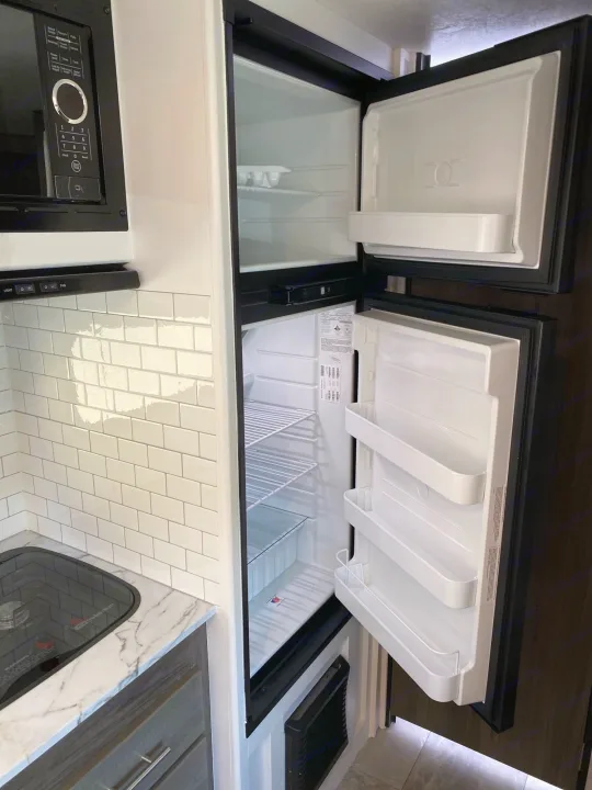 Bright RV rental kitchen with an open, empty refrigerator/freezer, microwave, and cooktop, showcasing its amenities.. Dutchmen Kodiak 2021
