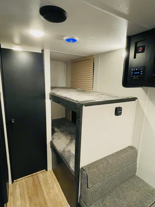 RV rental interior: Bunk beds, a small gray couch, black door, and ceiling lights create a compact, functional space.. Forest River Wolf Den 16EV 2021
