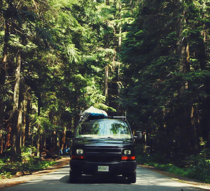 A black RV rental van with a kayak on its roof is parked on a sun-dappled road in a dense forest.. Chevrolet Express 2004