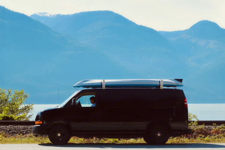 Black cargo van with kayak, parked by train tracks & lake, backed by majestic mountains. Perfect for an RV rental adventure!. Chevrolet Express 2004