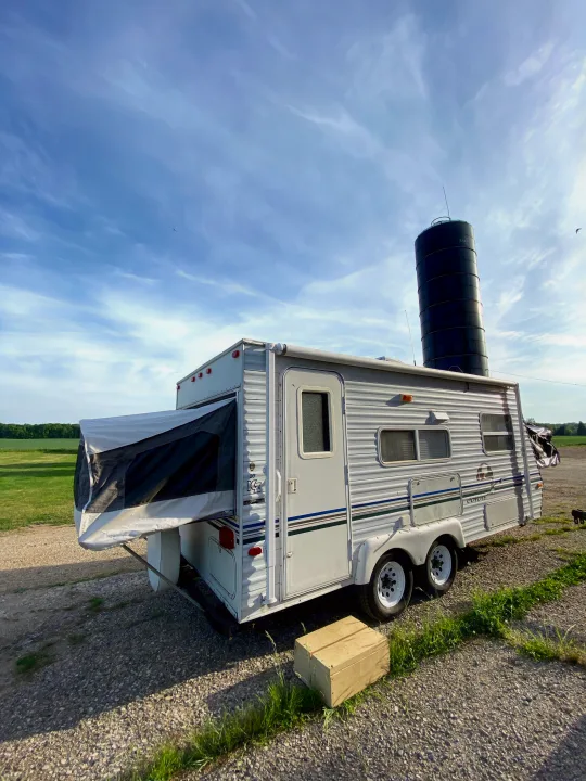 Explore the outdoors with this RV rental! A white travel trailer with an extended pop-out is parked on gravel near a silo and fields.. Sportsmen Coyote 2001