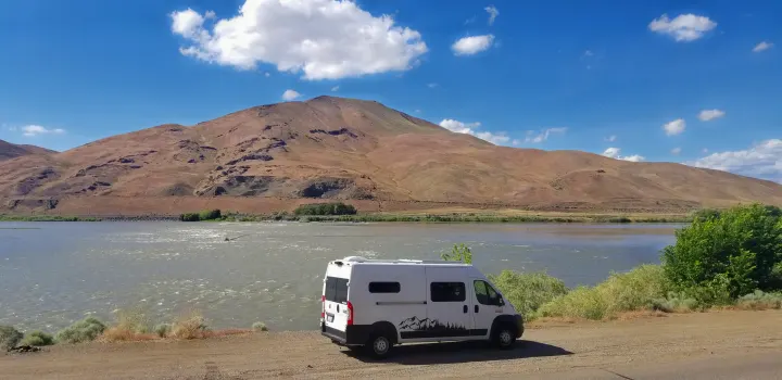 A white camper van rv rental parked on a dirt road by a wide river, with dry brown mountains under a blue sky.. Dodge Ram Promaster 2500 2019