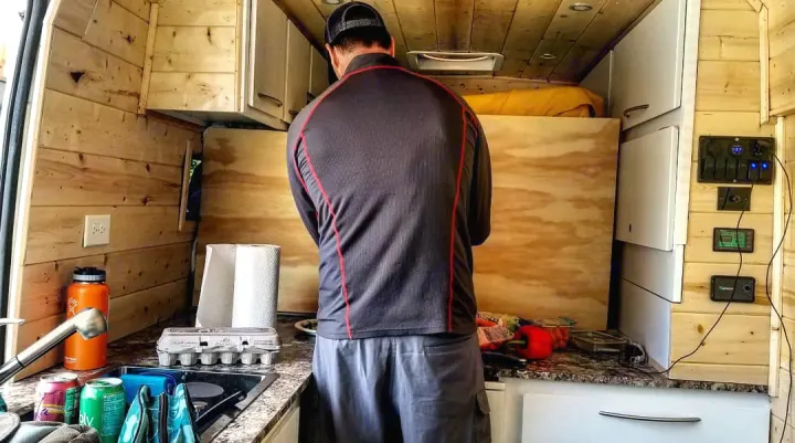 An RV rental interior with a person at the kitchen counter, showing wooden walls, counter, controls, and bedding.. Dodge Ram Promaster 2500 2019