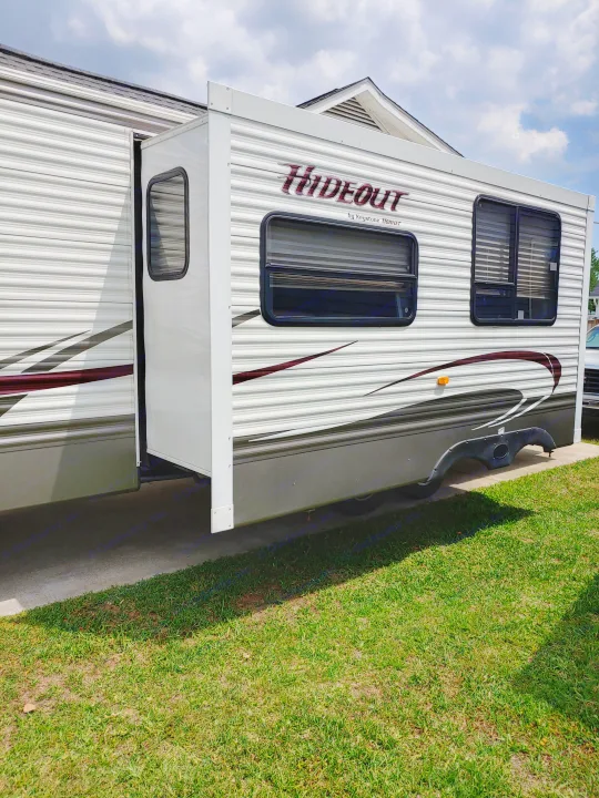 This white Hideout travel trailer RV rental features an extended slide-out, parked on concrete beside green grass.. Keystone RV Hideout 2014