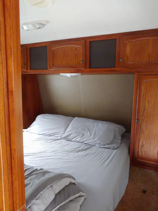 An RV rental bedroom featuring a neat bed with light grey linens, overhead wooden cabinets, and a compact design.. Keystone RV Hideout 2014