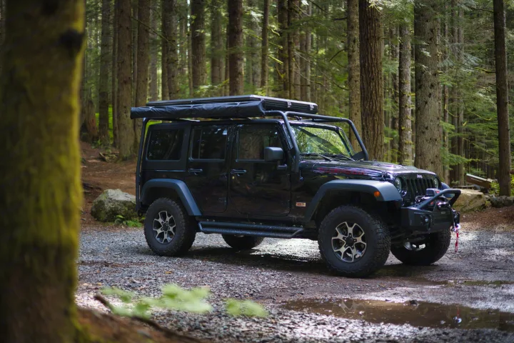 A black Jeep Wrangler with rooftop tent and awning is parked in a lush forest, offering an adventurous alternative to an RV rental.. Jeep Wrangler Unlimited Rubicon 4x4 2013