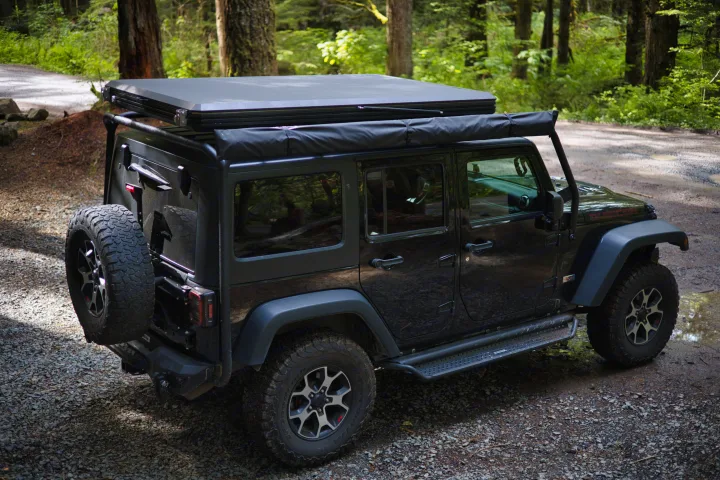 Black Jeep Wrangler with rooftop tent and awning, parked on a forest road. An ideal RV rental for overlanding adventures.. Jeep Wrangler Unlimited Rubicon 4x4 2013