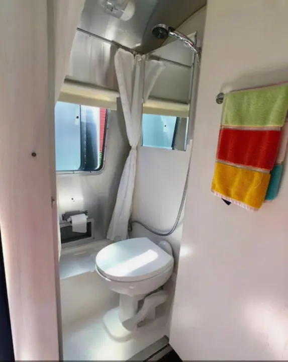 Inside an RV rental: a bright bathroom with toilet, shower, window, curtain, and striped towels hanging.. Airstream Sport 2018