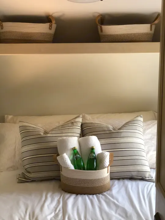 Cozy RV rental bed setup with striped pillows, welcome basket, rolled towels, and Perrier water bottles.. Winnebago Minnie Winnie 2018