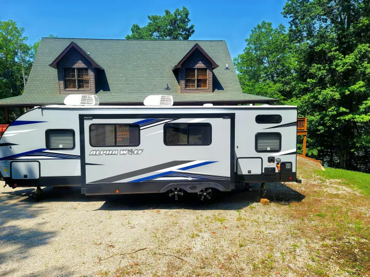 Experience nature with this modern rv rental. A gray Alpha Wolf travel trailer parked on gravel, backed by a house and green trees.. Forest River Cherokee Alpha Wolf 2021