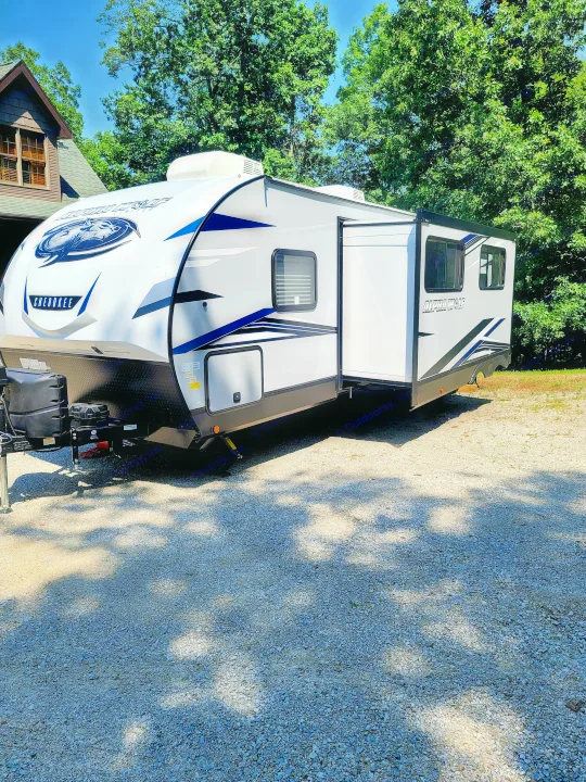 White Cherokee Alpha Wolf RV rental with an extended slide-out on a gravel lot, surrounded by green trees and a house.. Forest River Cherokee Alpha Wolf 2021