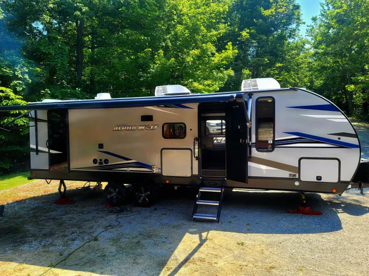 An Alpha Wolf RV rental is set up at a sunny, tree-lined campsite. Its awning is extended and steps are down, ready for guests.. Forest River Cherokee Alpha Wolf 2021