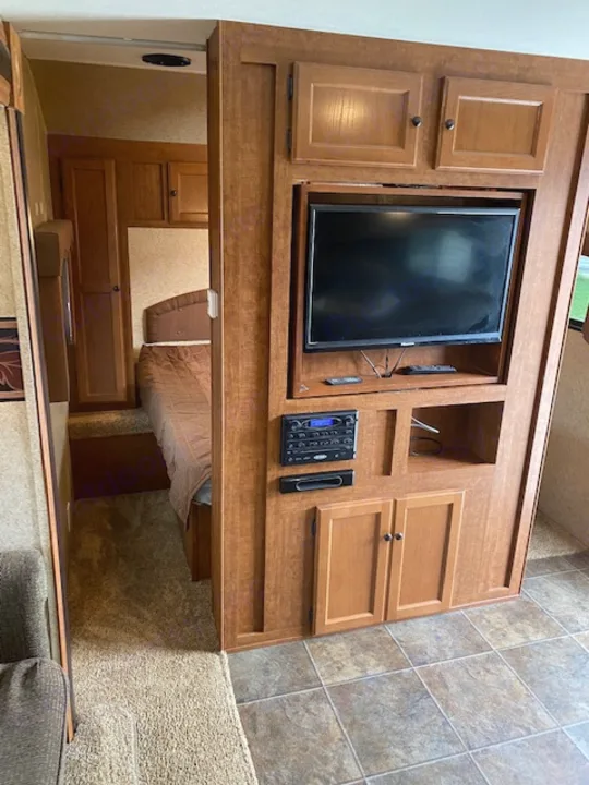 RV rental bedroom featuring a built-in entertainment center with a TV, control panel, and cozy wooden cabinetry.. Skyline NOMAD Joey Select 292 BHS  Double Wide Bunk 2014