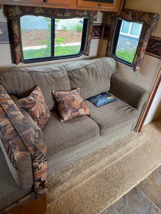 RV rental interior with a plush sofa, patterned pillows, and bright windows, showing a comfortable living area.. Skyline NOMAD Joey Select 292 BHS  Double Wide Bunk 2014