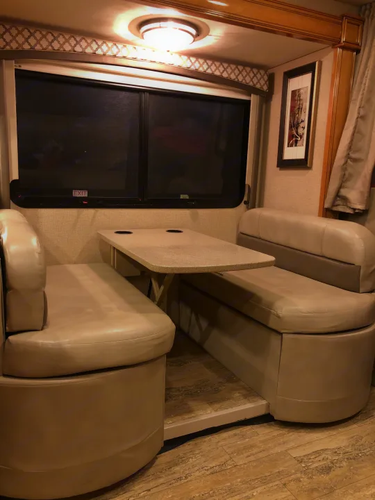 Cozy RV rental dinette with beige cushioned benches, a light table, and window. Ideal for dining and relaxing on the road.. Thor Chateau 2017