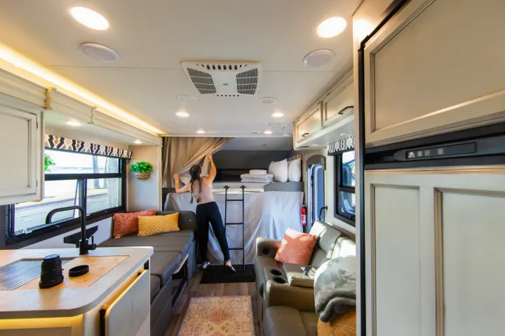 Modern RV rental interior: A person arranges curtains by the bunk bed, showcasing the kitchen, lounge, and amenities.. Jayco Greyhawk 2021