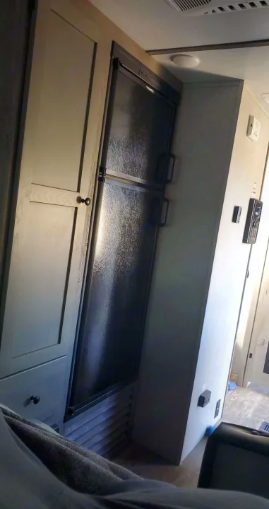 An RV rental interior features a dark Dometic refrigerator, adjacent light wood cabinets, and wall-mounted controls.. Keystone Outback 2020