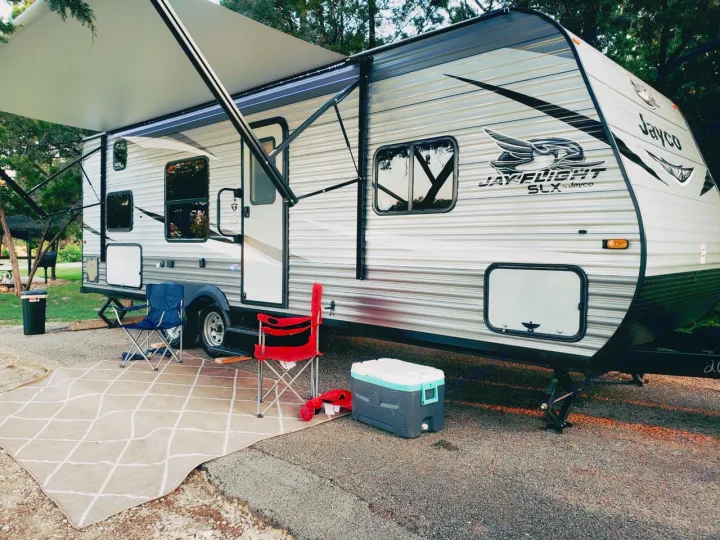 A large Jayco rv rental is set up at a shaded campsite, featuring an extended awning, outdoor rug, cooler, and two chairs.. Jayco Jay Flight SLX 2021