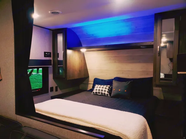 Inside a modern RV rental bedroom featuring a bed with blue accent lighting, wood walls, and a window overlooking grass.. Jayco Jay Flight SLX 2021