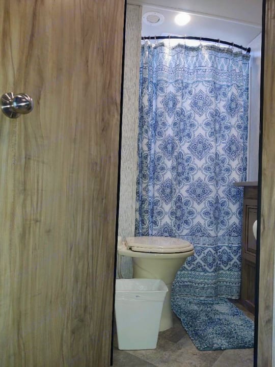 RV rental bathroom with a compact toilet, blue patterned shower curtain, and matching floor mat.. Palomino Solaire 243bhs 2021