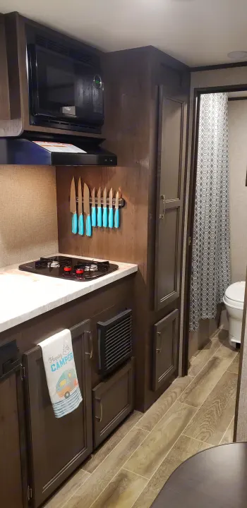 An RV rental's compact kitchen with a microwave, stove, and knife set. Wood-look flooring leads to a small bathroom.. Jayco Jay Flight 2021