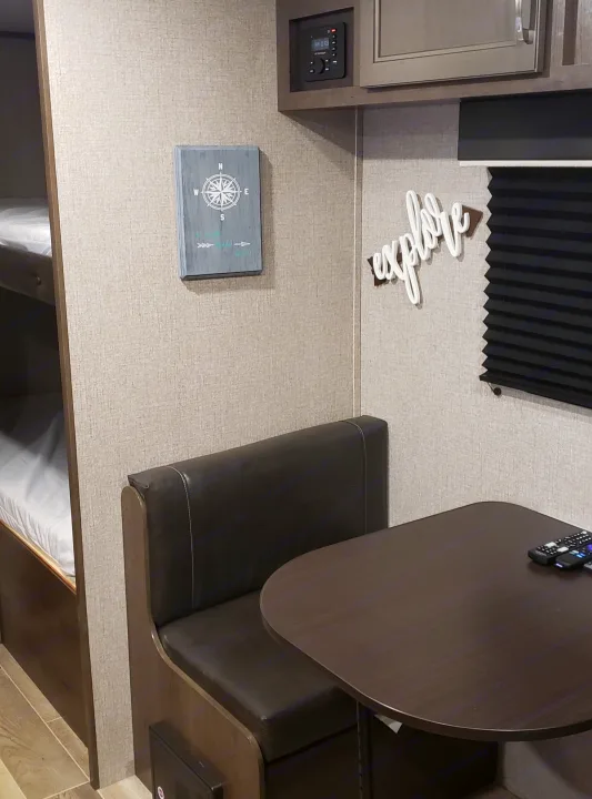 Inside an RV rental: a comfortable dinette with a dark table, bunk beds, and decorative wall signs like 'explore' and a compass.. Jayco Jay Flight 2021