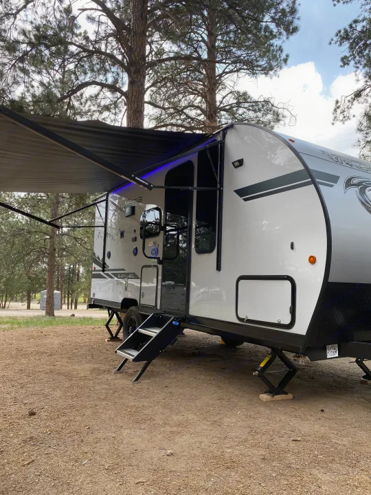 A modern travel trailer RV rental is set up on a rustic dirt campsite, featuring an extended awning with blue lights.. Forest River Cherokee Wolf Pup 16PFBL 2021