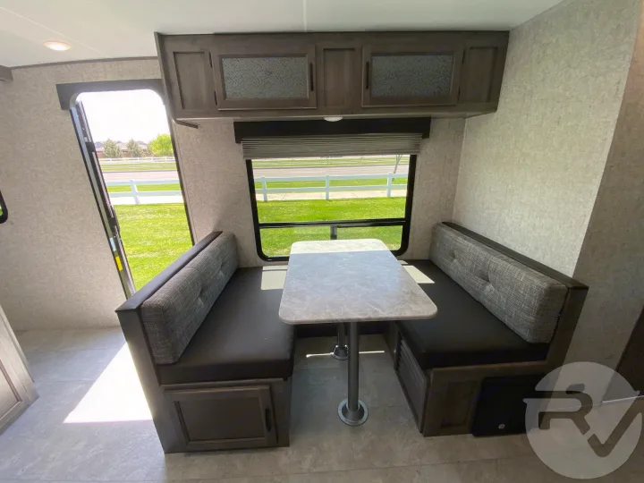 Bright RV rental interior showing a dinette with bench seating, a table, and overhead cabinets. Window view of outdoors.. Coachmen Apex 2021