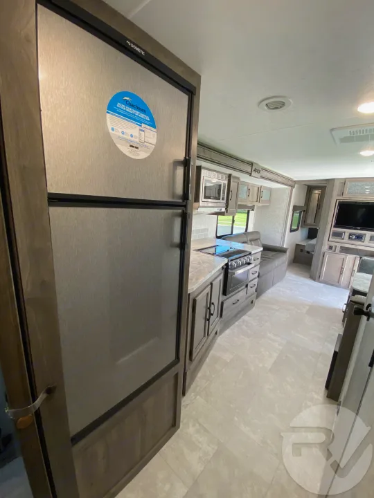 Inside a modern RV rental: a Dometic refrigerator anchors the compact kitchen with oven & microwave. Cozy living area beyond.. Coachmen Apex 2021