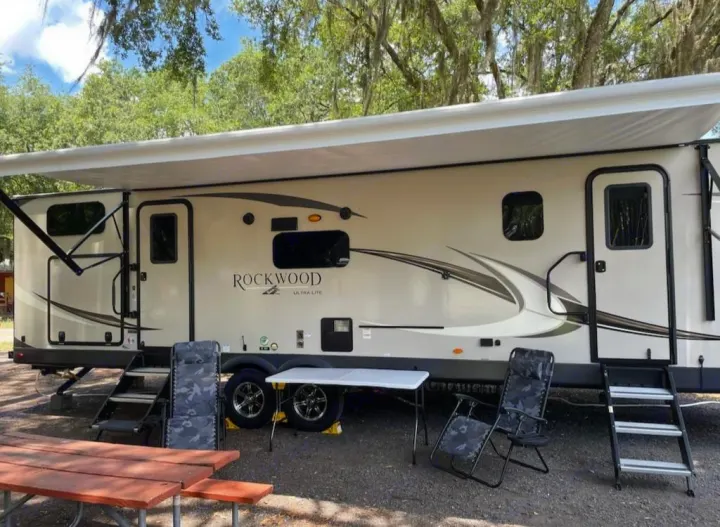 Cream-colored travel trailer RV rental with extended awning, outdoor seating, and table for a relaxing camping setup.. Forest River Rockwood Ultra Lite 2021
